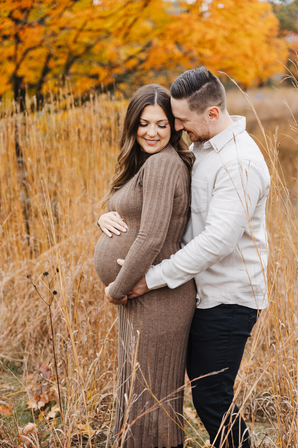 Expecting husband and wife embracing in the tall grasses of High Park. Husband is hugging wife from behind.