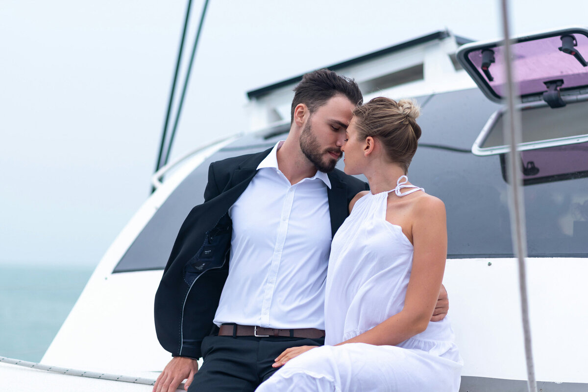 man in a black suit and white shirt sits close to a woman in a white dress on a yacht, embracing and about to kiss