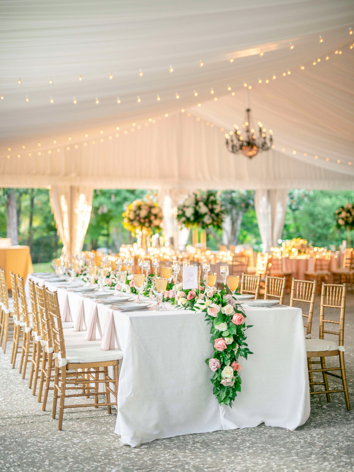 An elegant outdoor wedding reception with long tables draped in white linens, decorated with pink and white roses. Gold chairs surround the tables, and string lights hang from the ceiling. Tall floral arrangements are in the background.