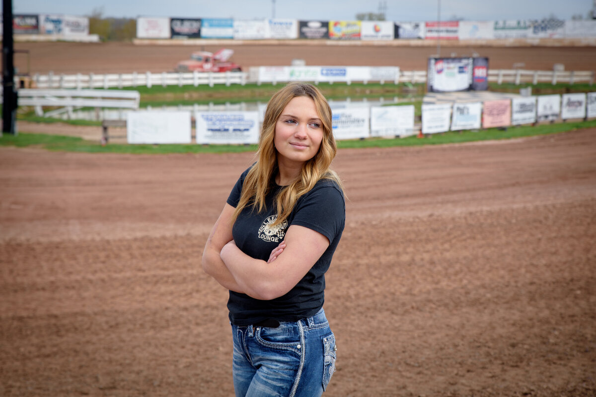 denmark high school girl is standing with arms crossed in front of dirt race track at 141 Raceway in Francis Creek, Wisconsin