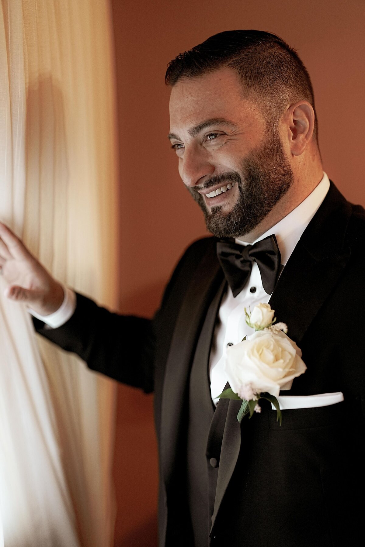 The groom smiles warmly as he gazes out of a window, captured in a moment of joy and contentment. This image highlights his cheerful demeanor and adds a touch of elegance and serenity to his wedding portraits.