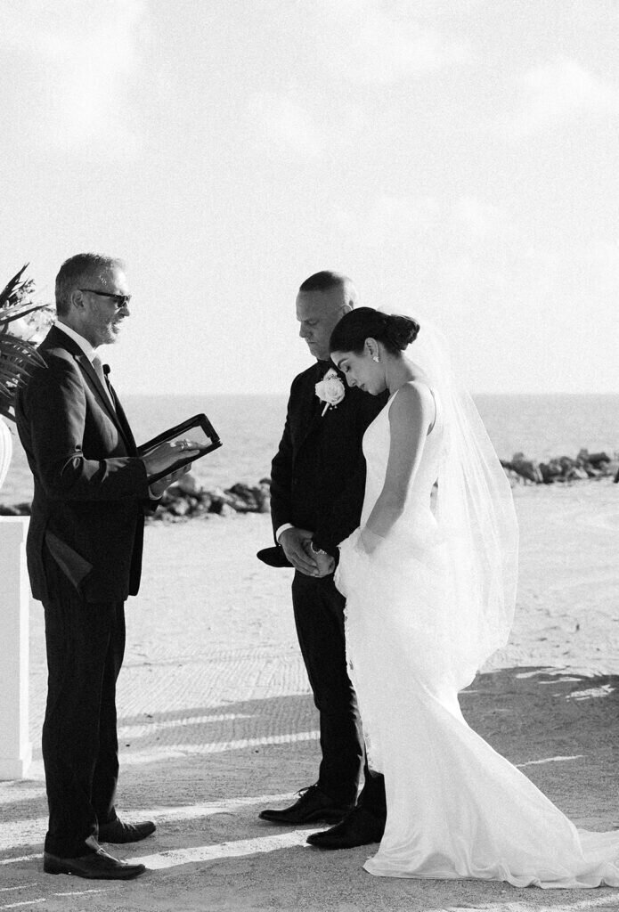 A heartfelt black-and-white moment as the bride and groom bow their heads in prayer during their beachfront wedding ceremony. Destination wedding photography by Claudia Amalia Photography.