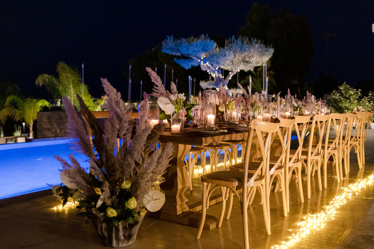 Naked wooden tables with cross back chairs are  adorned with rustic foliage set against a lit pool