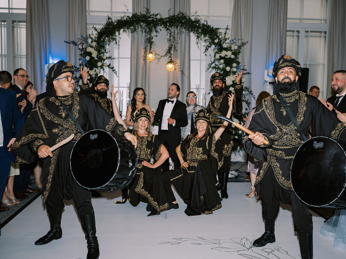 At a classic Calgary wedding, a group of people in traditional attire perform a dance with drums at an indoor event, surrounded by an audience. The background features large windows, curtains, and a decorative floral arch.