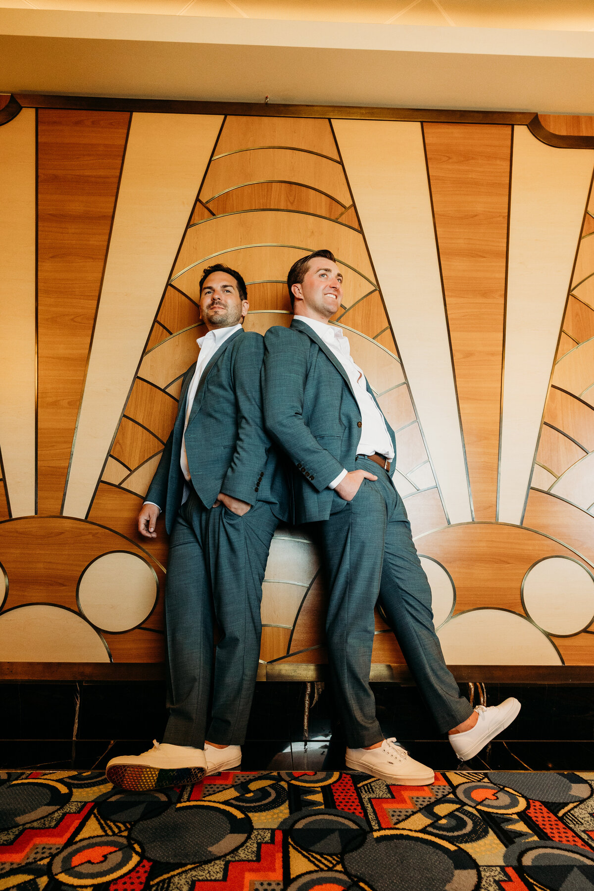 Two grooms pose back-to-back in front of an art deco wall on the Disney Fantasy during their cruise wedding portrait session.