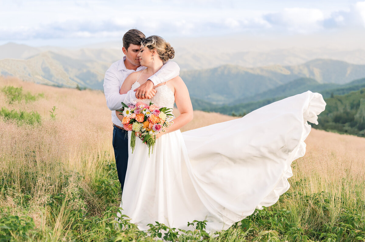 A bride and groom sharing sunset portraits on The Blue Ridge Parkway at sunset as the wind is blowing the bride's dress up in the air as they are enjoying their Raleigh wedding photography session by JoLynn Photography