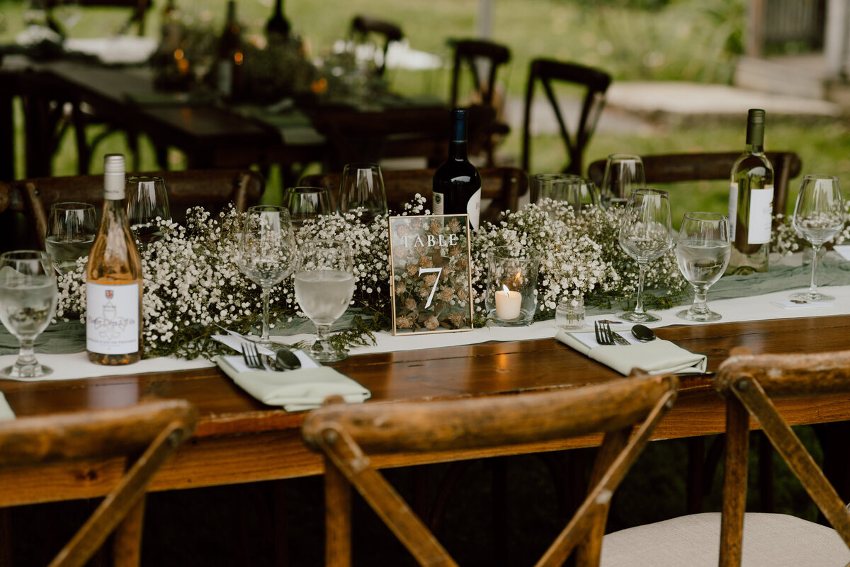 Rustic wedding reception table adorned with baby's breath floral arrangements, candles, and wine bottles, set for an intimate outdoor celebration. The wooden table is beautifully set with glassware, cutlery, and a table number card, creating a charming and inviting atmosphere.