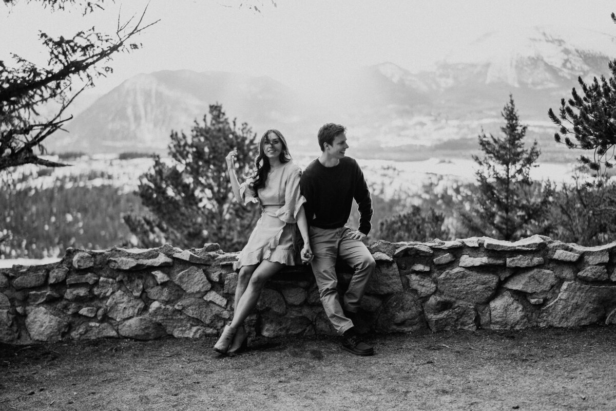 Engaged couple sits on stone wall holding hands during Breckenridge engagement session.