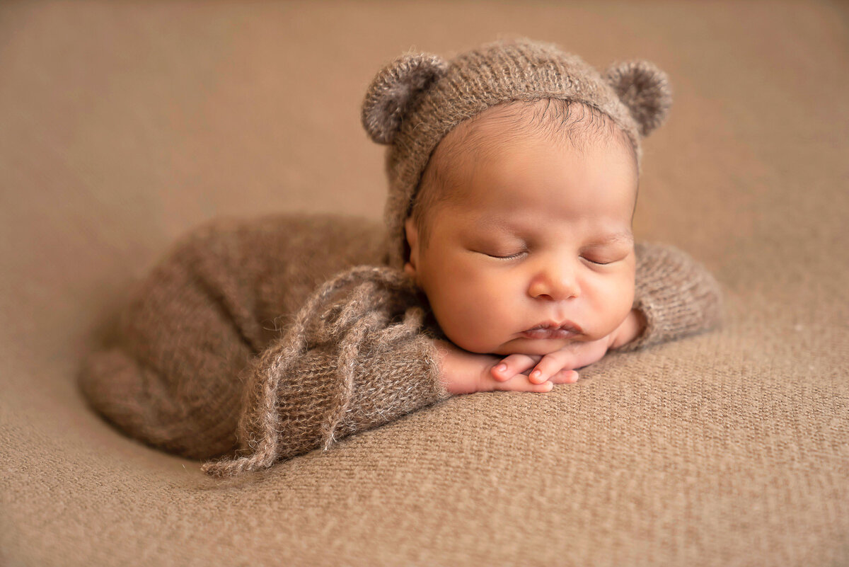 Baby boy in knit teddy bear bonnet is sleeping with his hands folded under his chin for a newborn shoot in Bryn Mwar photography studio. Captured by Bryn Mwar Philadelphia and Main Line photographer Liza Nicole.