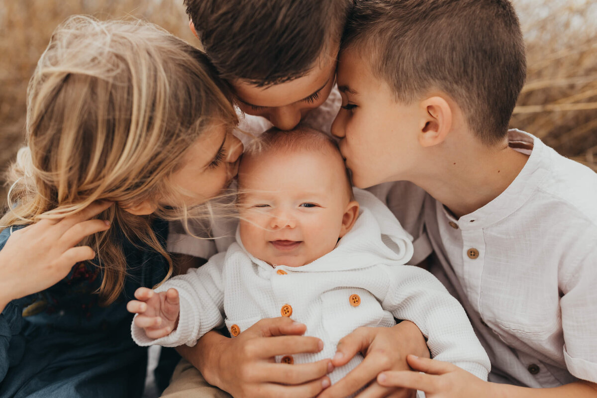 baby boy smiling while siblings kiss him on the head