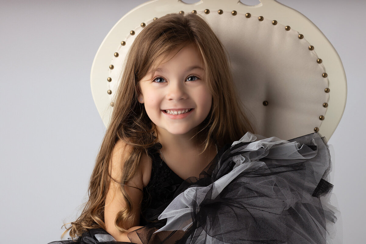 A close-up of a smiling young girl in a black dress with gray ruffles, sitting in a vintage chair.