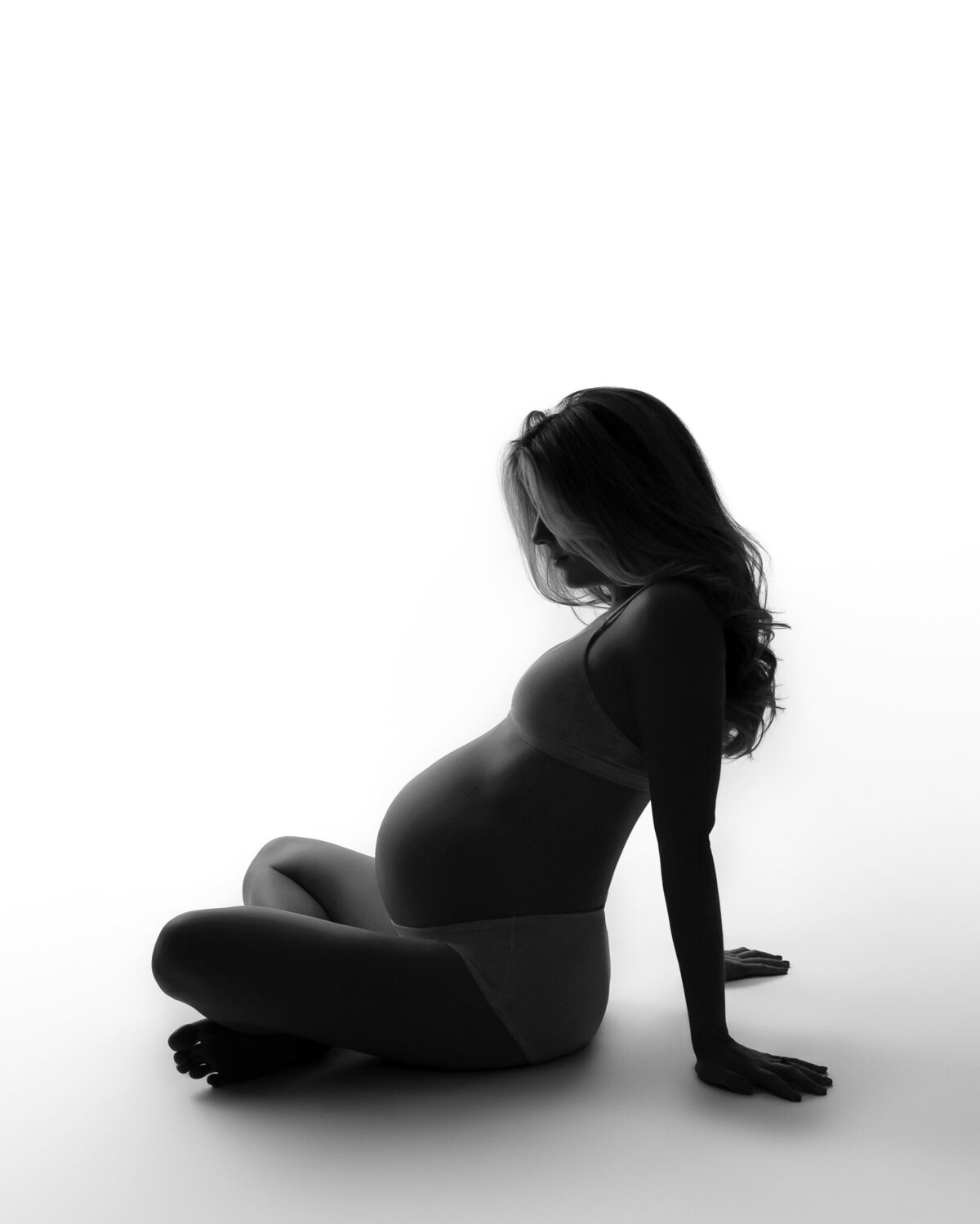 Clean and modern studio portrait of a pregnant woman sitting cross-legged in silhouette, lit softly from behind.