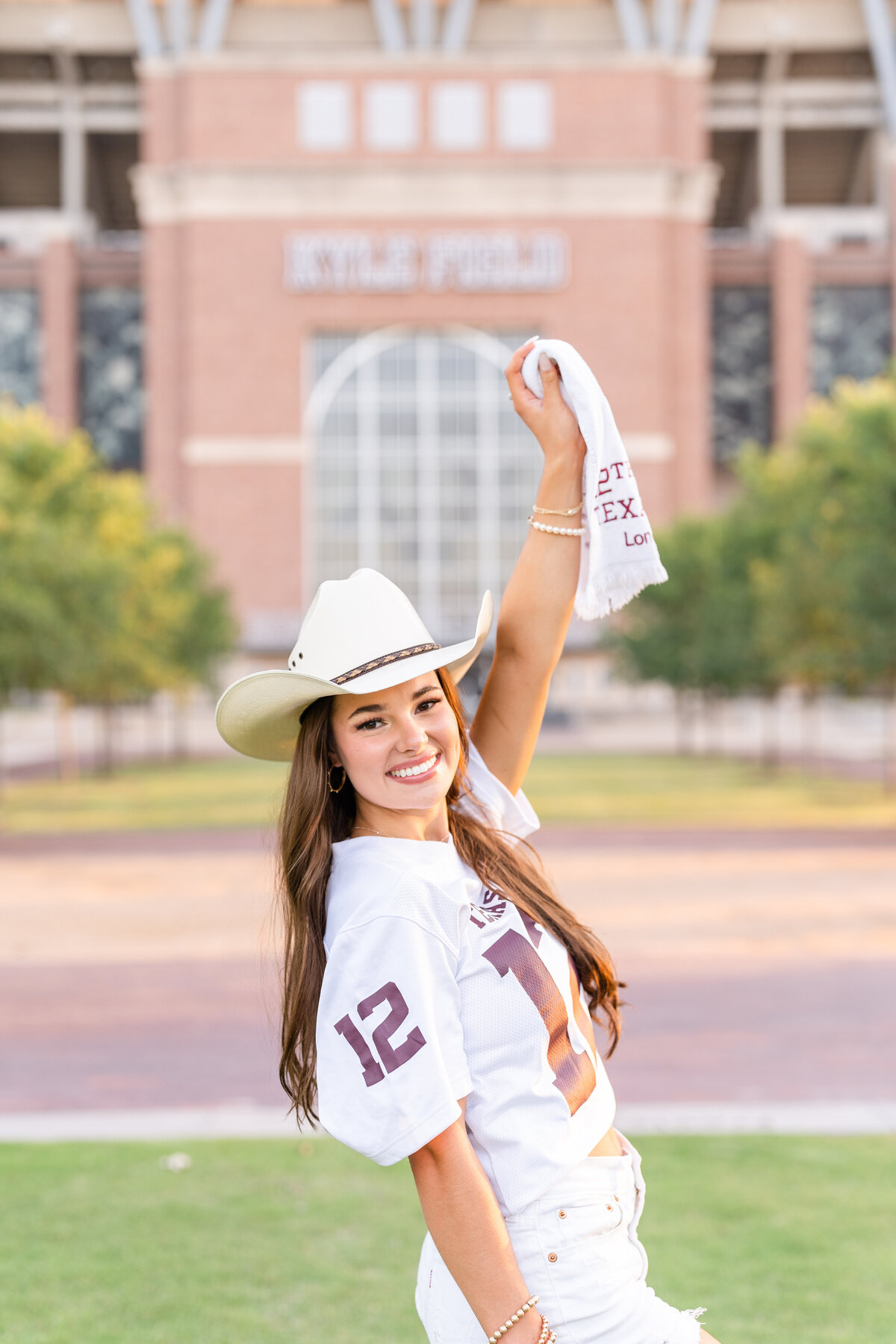 Texas A&M senior girl wearing white jersey and cowboy hat smiling at camera and holding up 12th Man towel in front of Kyle Field at Aggie Park