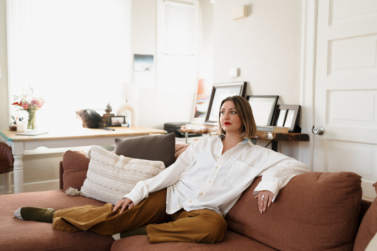 "A woman with a bob haircut and red lipstick sits casually on a brown sofa in a bright, cozy living room. She wears a white shirt and mustard-colored pants, with framed artwork and a work desk visible in the background."
