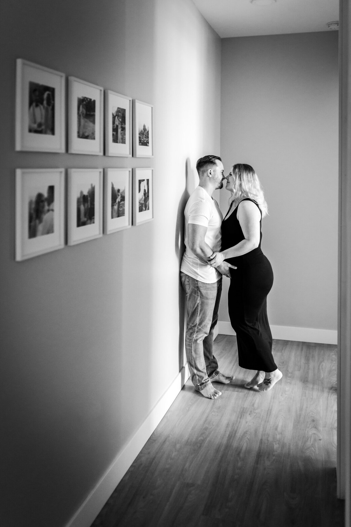 A couple stands in a narrow hallway, facing each other and holding hands, about to share a kiss. The walls are decorated with a series of framed photos.