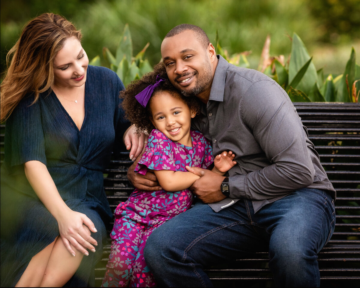 Caucasian mom with African American father and biracial daughter are sitting on a bench in Audubon Park.  They are laughing and hugging each other.  The mom is wearing a blue dress, the dad is wearing gray shirt and jeans, and the 4 year old daughter is wearing a purple floral jumpsuit.  The daughter has natural hair.