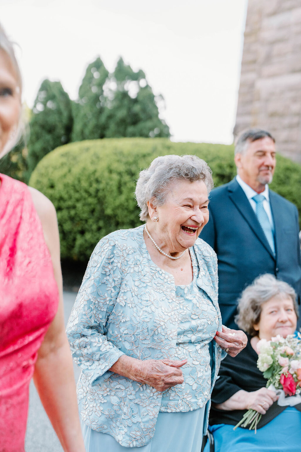 Wedding Photographer RI | Elderly woman in a light blue floral dress laughing joyfully outdoors, surrounded by people dressed for a wedding. Greenery and trees in the background.