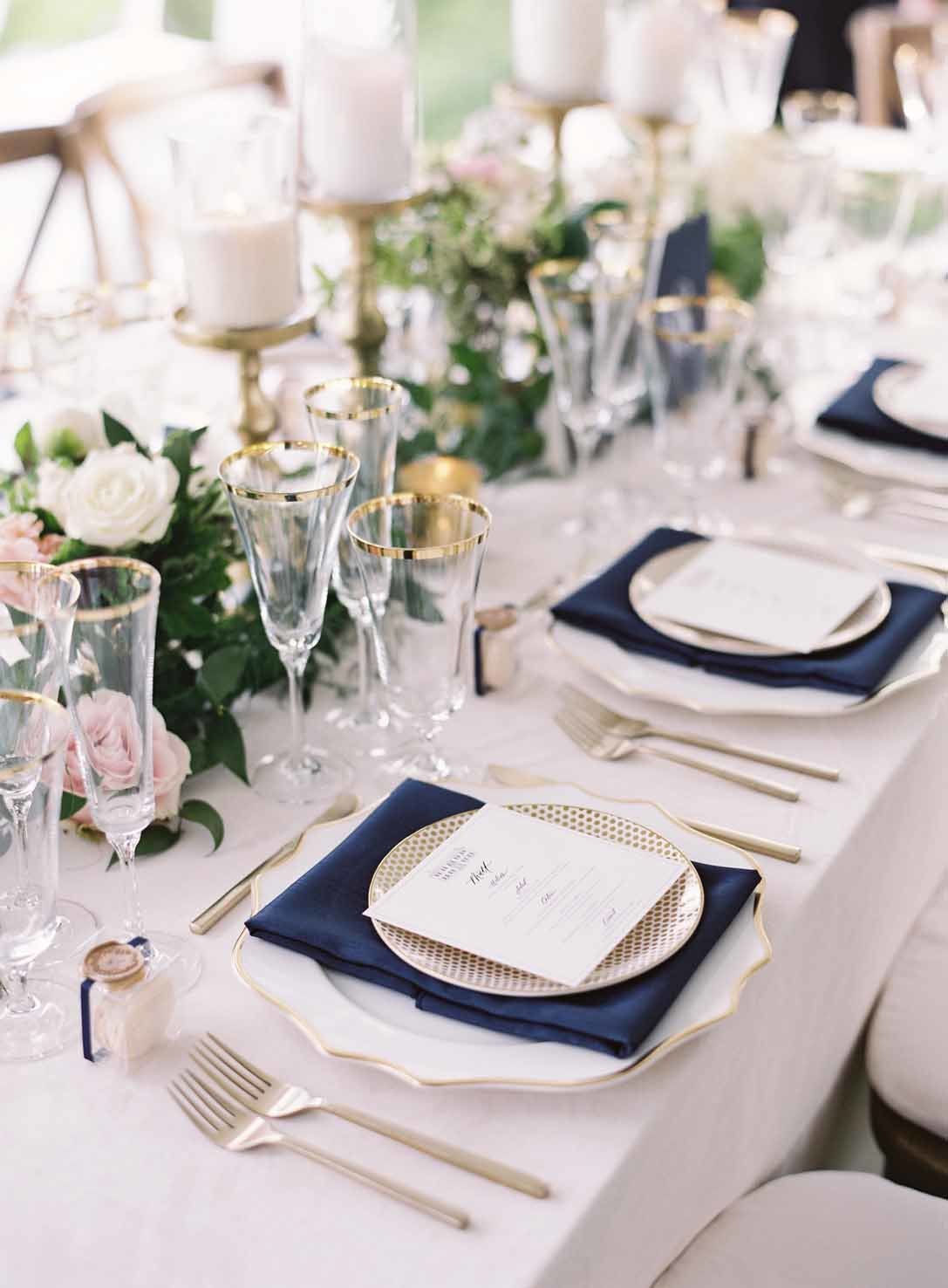 long reception table lined with flowers and candles, gold flatware, navy blue napkins, and gold rimmed plates
