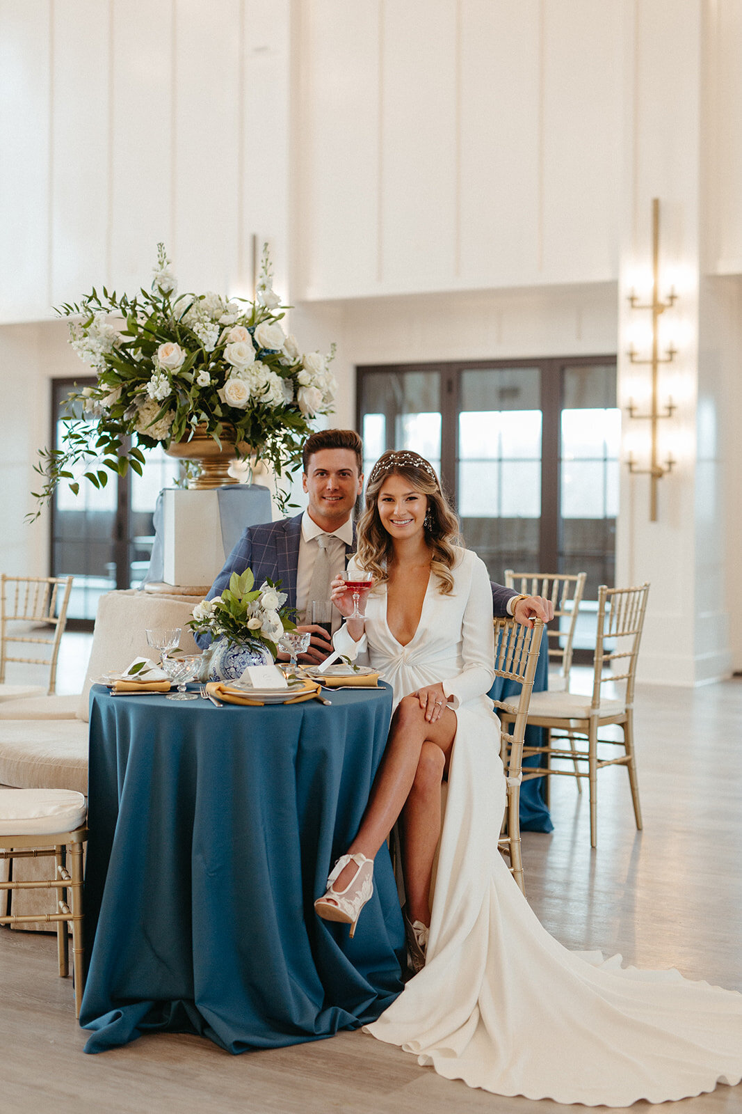 Bride and groom in a blue suit and white wedding gown seated at a banquet table holding a cocktail.
