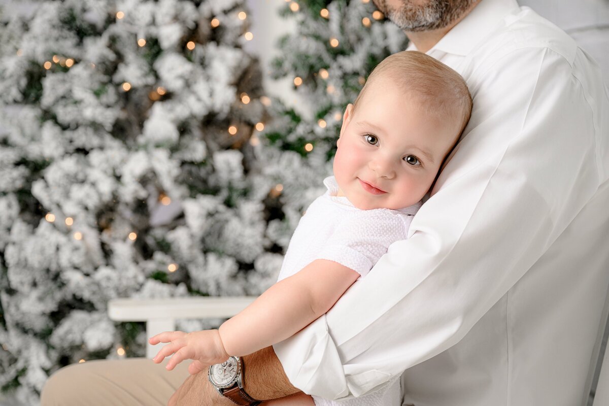 little boy dressed in white leans back against his father's shoulder in white christmas  set up