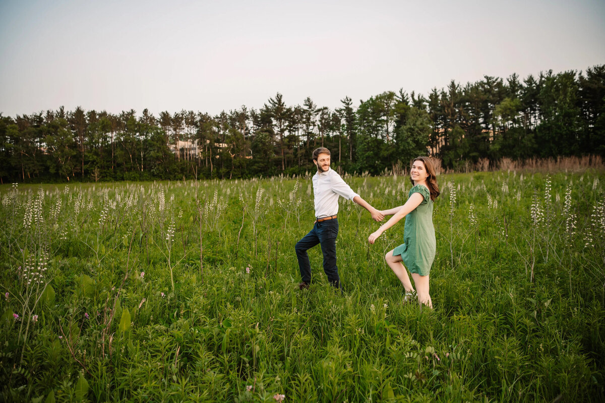 man and woman walk in field holding hands while woman looks a the camera and man looks at woman