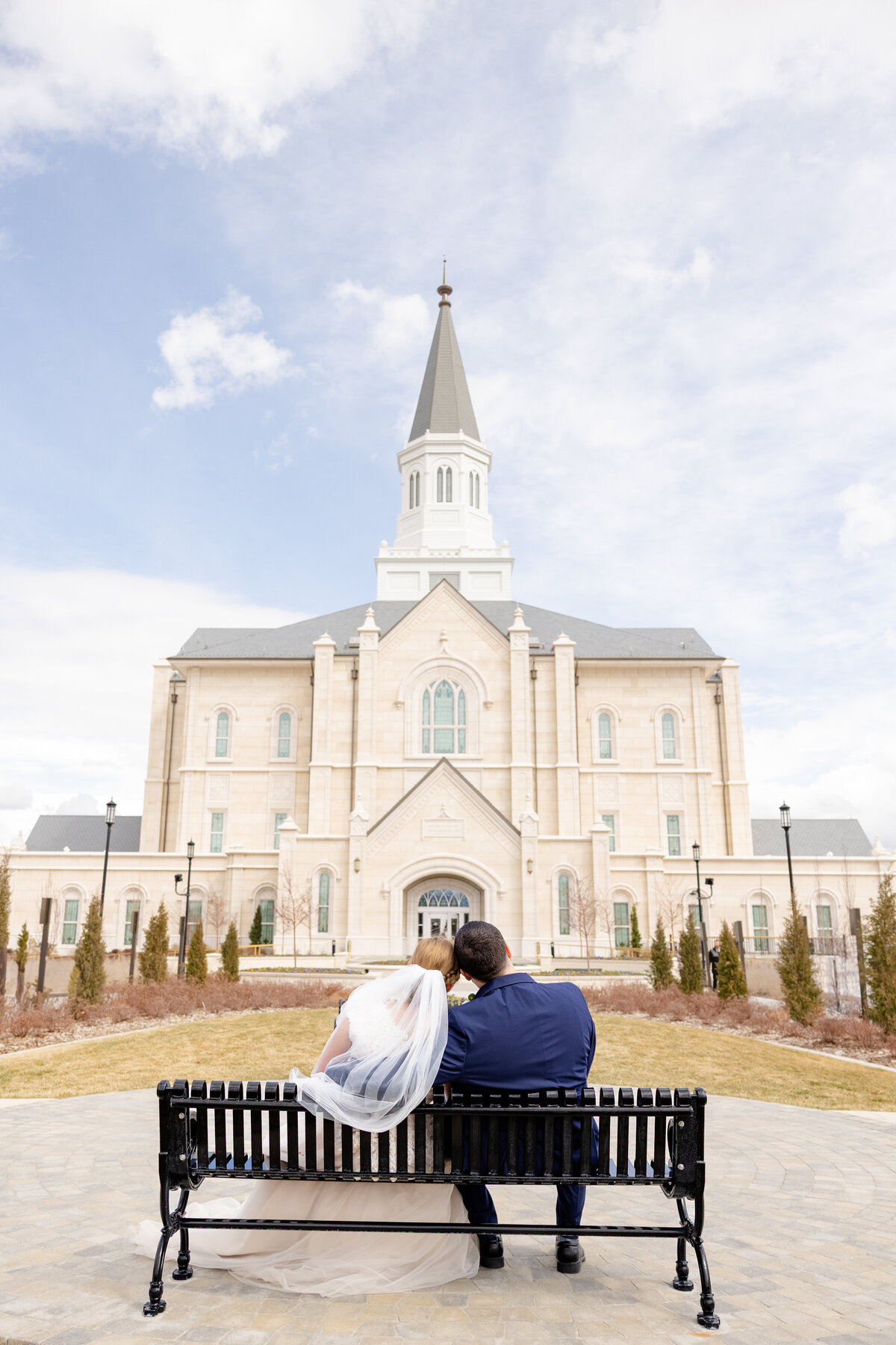 Bride and groom sit closely on a black bench, gazing at the Taylorsville Utah Temple’s stunning architecture on a bright wedding day.