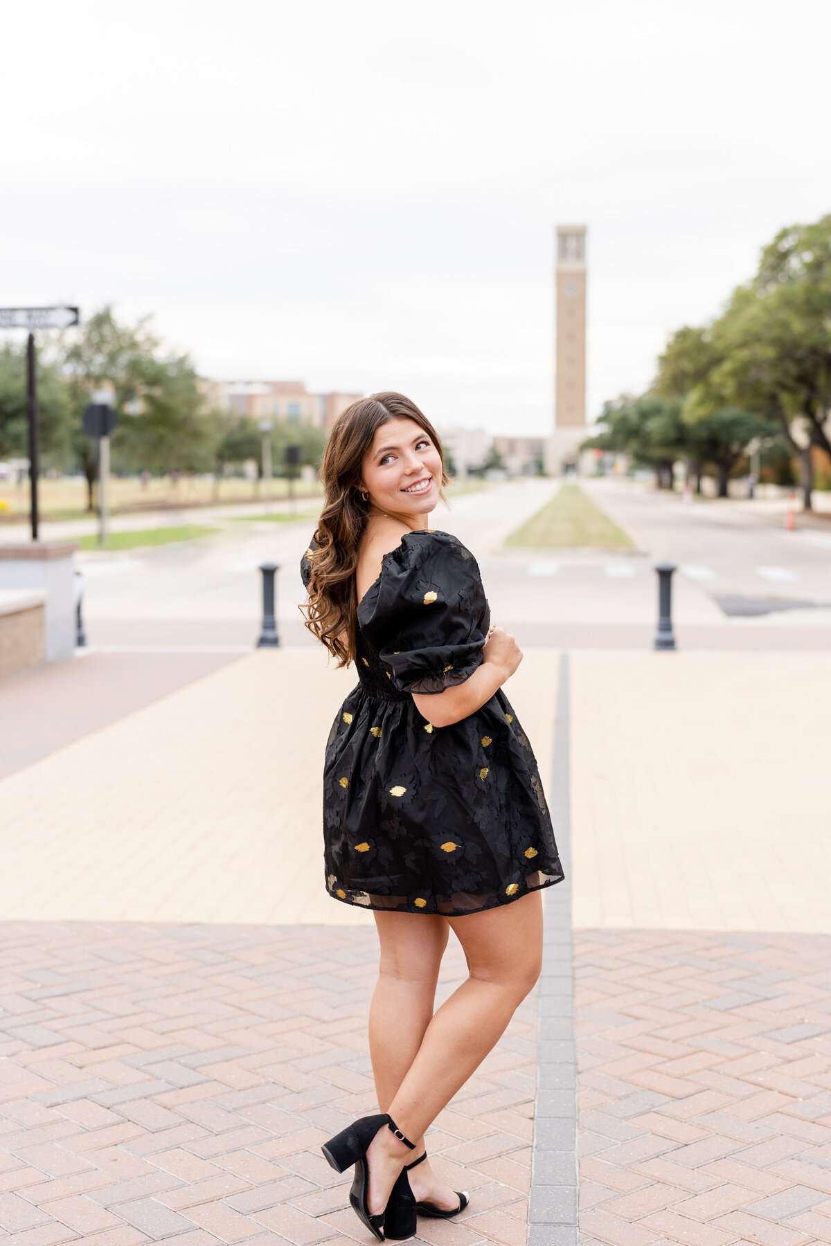 Texas A&M senior girl wearing a black dress and looking over shoulder smiling with Bell Tower in the background