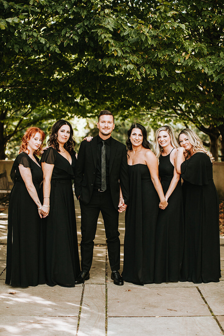 The wedding party poses outside wearing black gowns and a black suit, with large trees in the background.