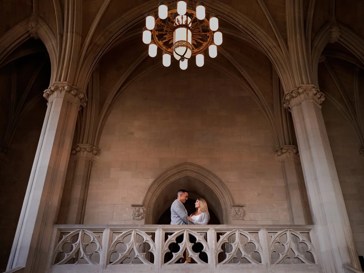 Couple sharing an intimate moment on a balcony under an elegant chandelier within a gothic-style architecture