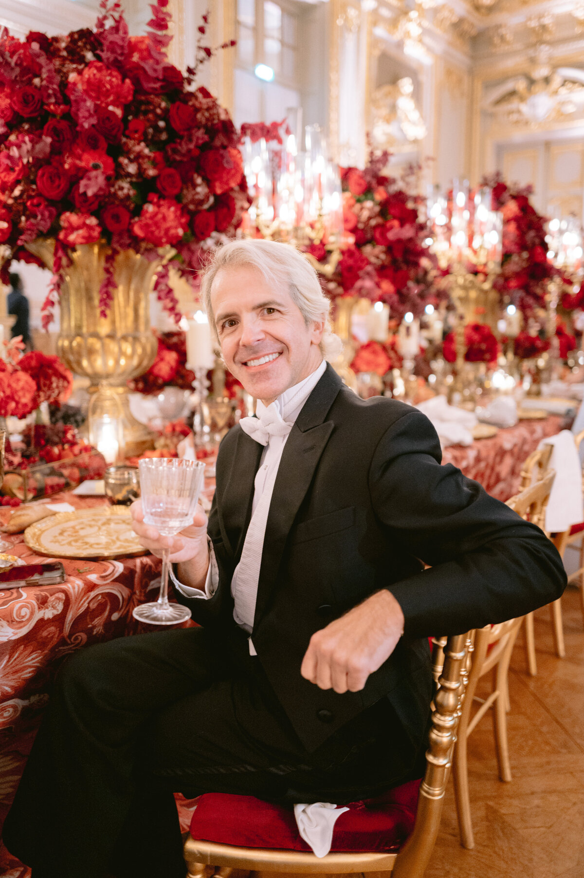 The photo was captured at the luxurious Shangri La Hotel & Ritz Paris Hotel in Paris, France, by the talented photographer Matteo Coltro, with videography services provided by Zen Film Works. In the elegant scene, a person in a formal black tuxedo sits at an exquisitely decorated table adorned with a lavish red and gold theme, holding a wine glass. The table is beautifully complemented by ornate floral arrangements and softly lit candles, all within an opulent room featuring intricate architectural details and sparkling chandeliers.