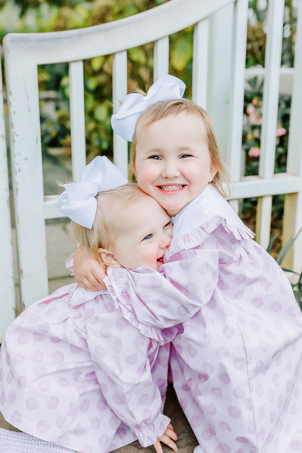 Two sisters share a joyful hug in matching lavender outfits with white bows.