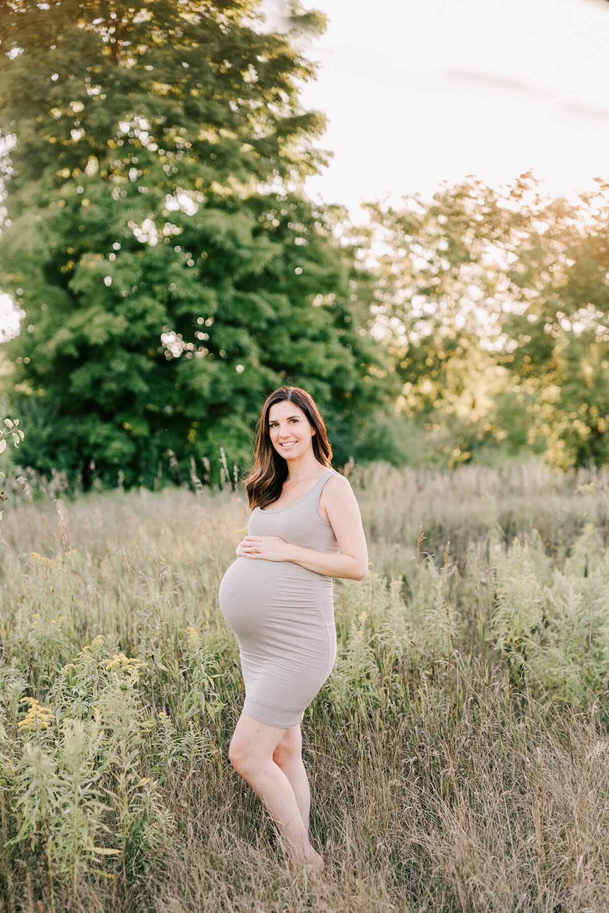 woman in taupe fitted dress stands in the field for her maternity session.