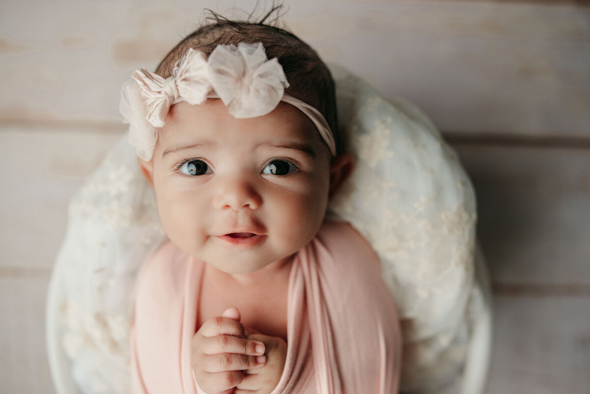 Fairfield CT New Haven CT studio Smiley bright eyed newborn baby girl with brown hair and brown eyes wrapped in pink swaddle, wearing a white bow headband and laying on white fabric