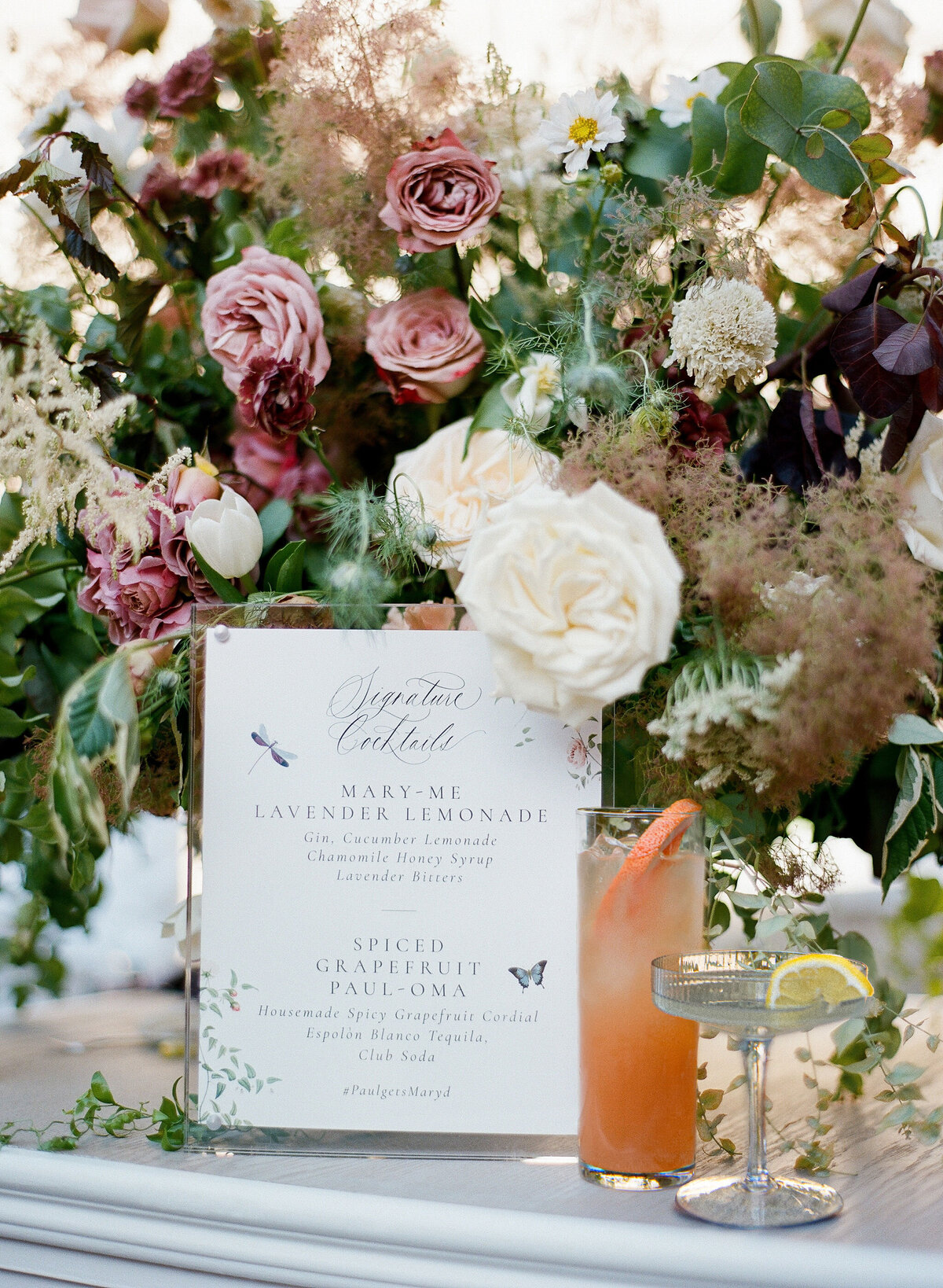 At a picturesque location in Washington D.C. stands the majestic Washington National Cathedral. The serene ambiance captured in the photograph by photographer Twah Dougherty perfectly frames a beautifully arranged floral display featuring roses, wildflowers, and greenery. Amongst the vibrant blooms, a menu card listing Signature Cocktails is elegantly placed. Adjacent to it, a glass of pink grapefruit drink garnished with a lime wedge adds a pop of color to the scene. The lush, garden-like setting in the background enhances the overall charm of this enchanting setting. Current Films as Videographers.