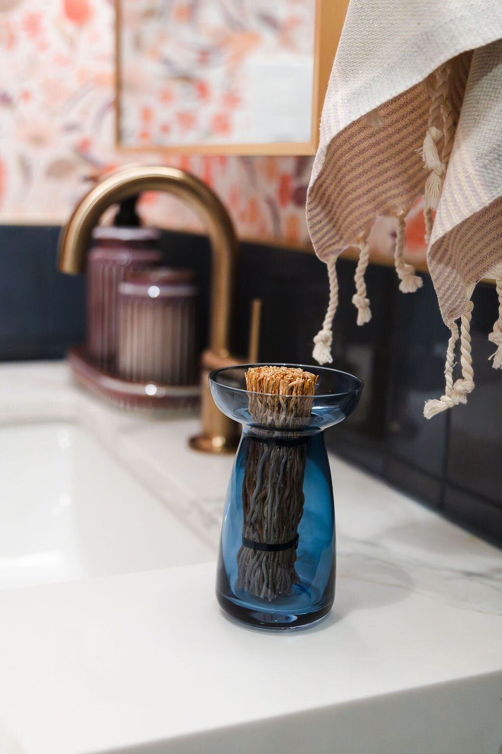 A close-up of a bathroom countertop featuring a white surface. A small, elegant blue glass vase with a pedestal base holds dried natural elements. A light-colored hand towel with thin stripes and tassels hangs from a fixture above the counter. The backsplash is made of dark blue horizontal tiles with a thin gold trim at the top. The wall above the tiles is covered with a vibrant floral patterned wallpaper in shades of pink, orange, and white. A portion of a wooden-framed mirror is visible in the upper left corner. Soft lighting illuminates the scene.