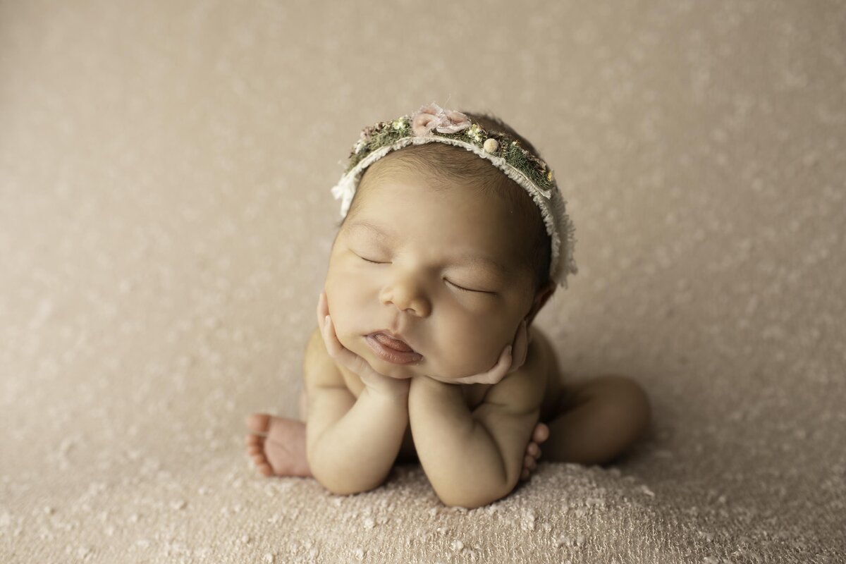 Newborn baby girl posed in the classic froggy position, her tiny hands gently supporting her chin, wearing a delicate headband. She rests peacefully on a soft backdrop, creating a sweet and timeless portrait