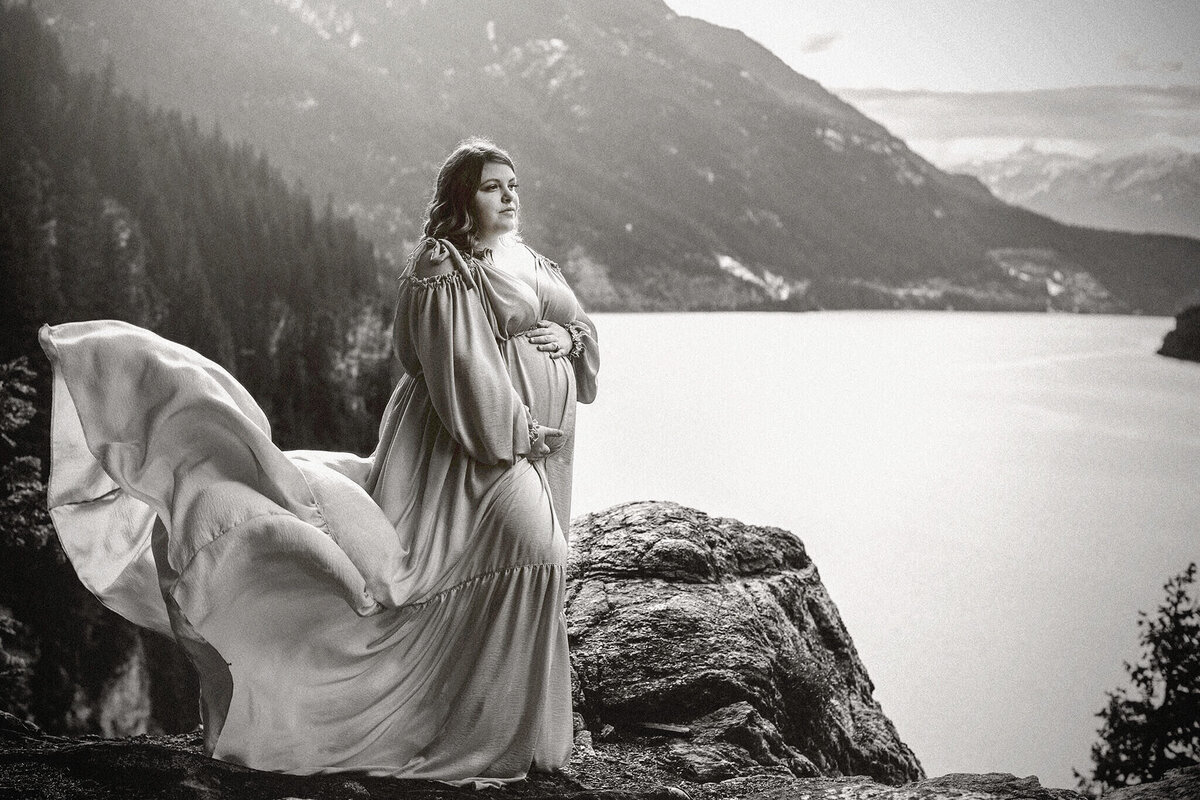 A pregnant women in a silk flowing gown stand on the edge of a cliff with a beautiful lake in the background. Photo taken in Revelstoke by Brittany Danielle Photography