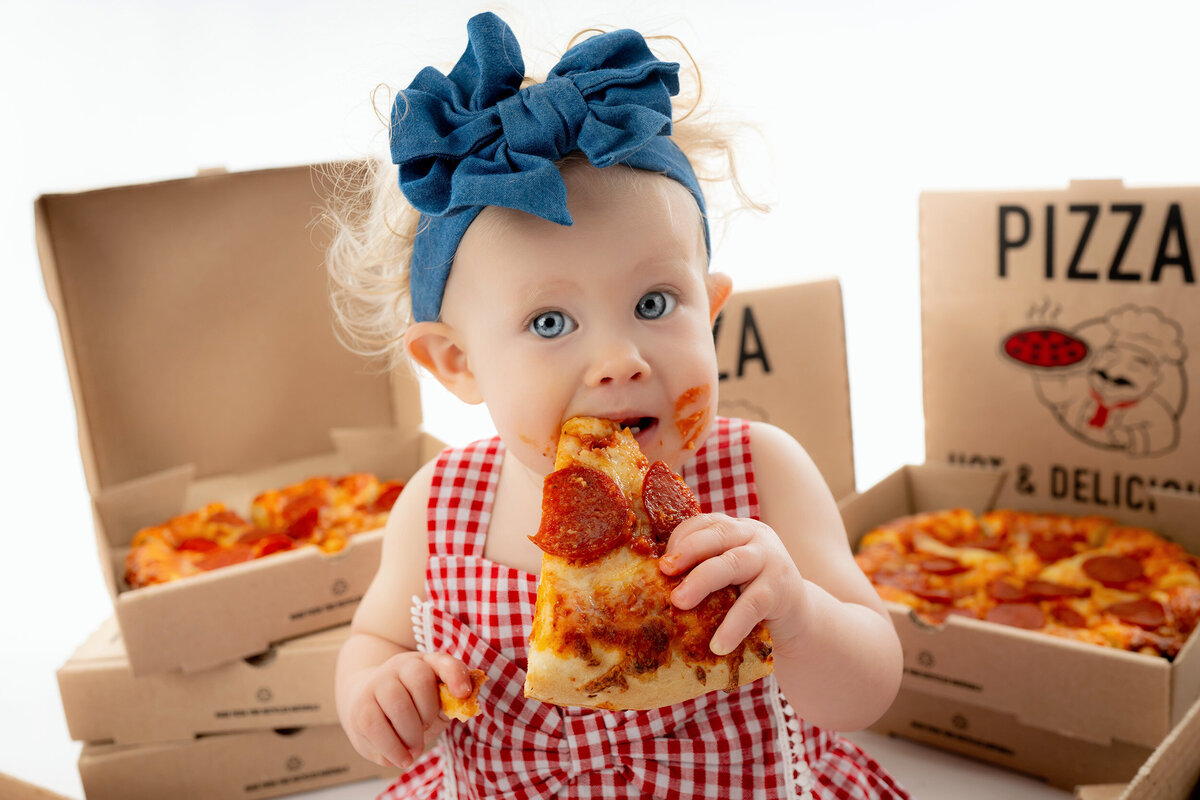 A toddler in a checkered outfit eating a slice of pizza with pizza boxes in the background.