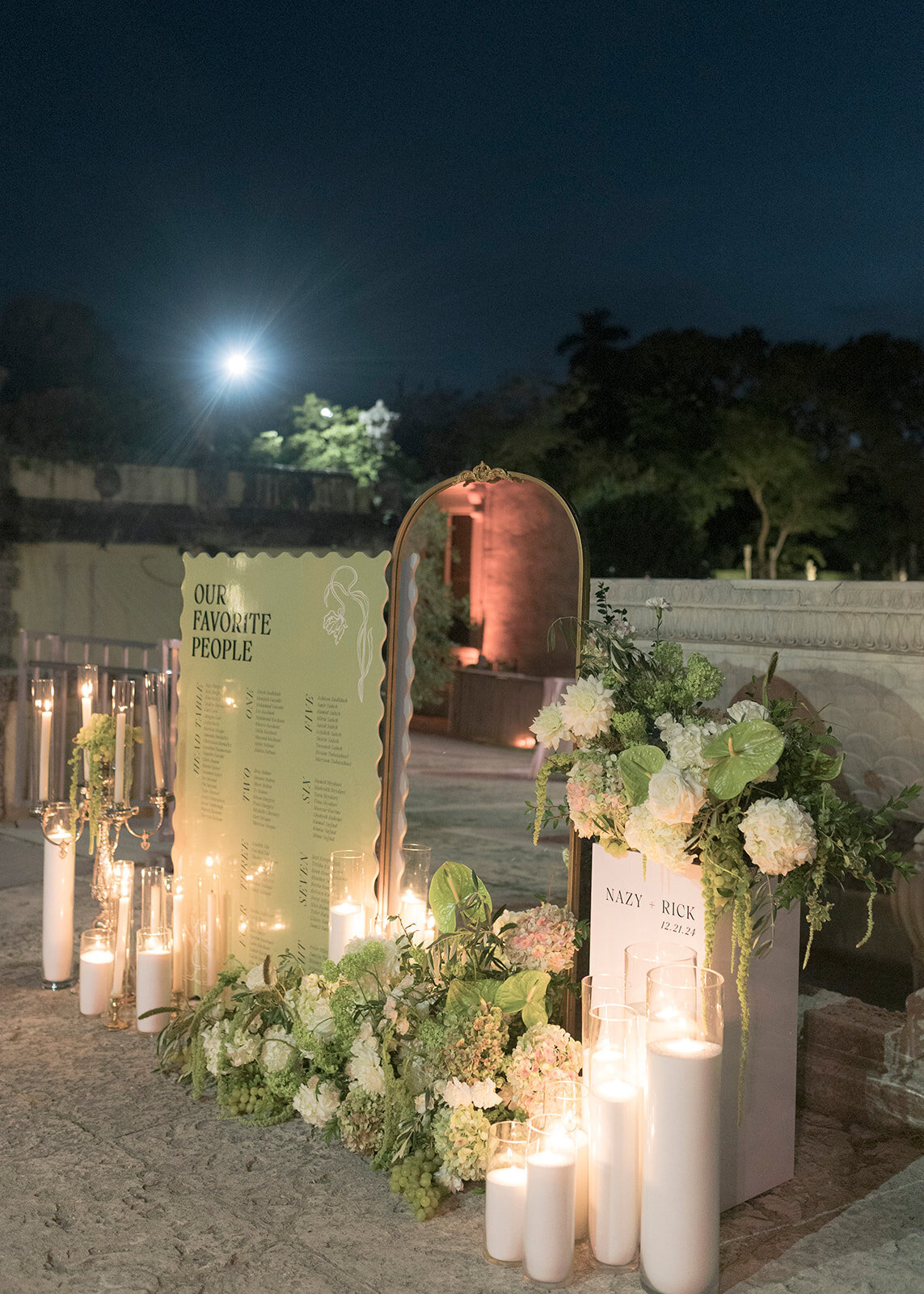 Stunning cocktail hour florals for wedding reception at Vizcaya Museum and Gardens in Miami, Florida. Lush floral meadows and candles decorate the escort display with dripping amaranthus and antique hydrangeas. Grazing table decorated with styled fruit and charcuterie for cocktail hour refreshments. Italian-inspired wedding design with heavy draping and green and white florals. Design by Rosemary & Finch Floral Design in Nashville, TN.