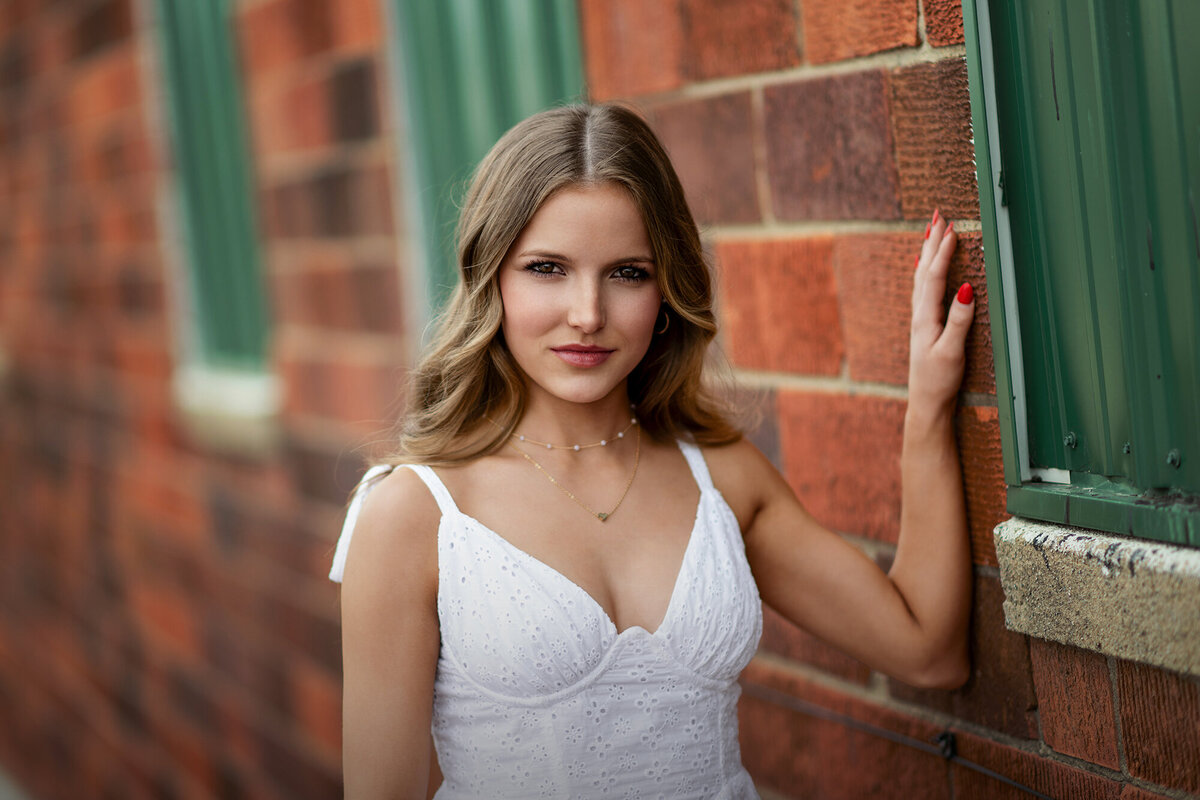 A Des Moines Senior Photography image of a high school senior in a white dress leans on a brick wall