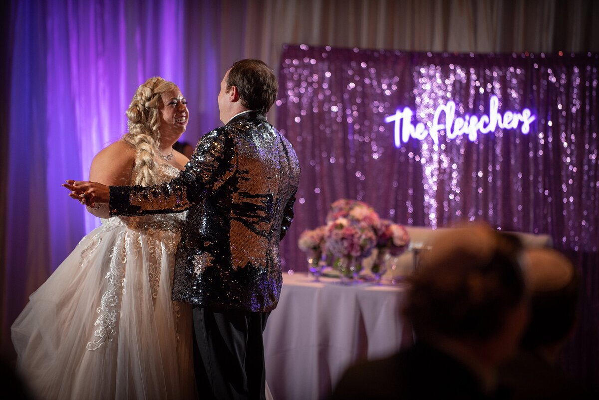 Bride wearing a flowing tulle wedding dress with a long braid dances with the groom who is wearing a purple sequined tuxedo jacket at their wedding at The Liff Center. The sweetheart table in the background has a light purple table cloth and a floral centerpiece in a silver footed bowl. Behind the sweetheart table is a purple sequined backdrop with a purple neon sign.