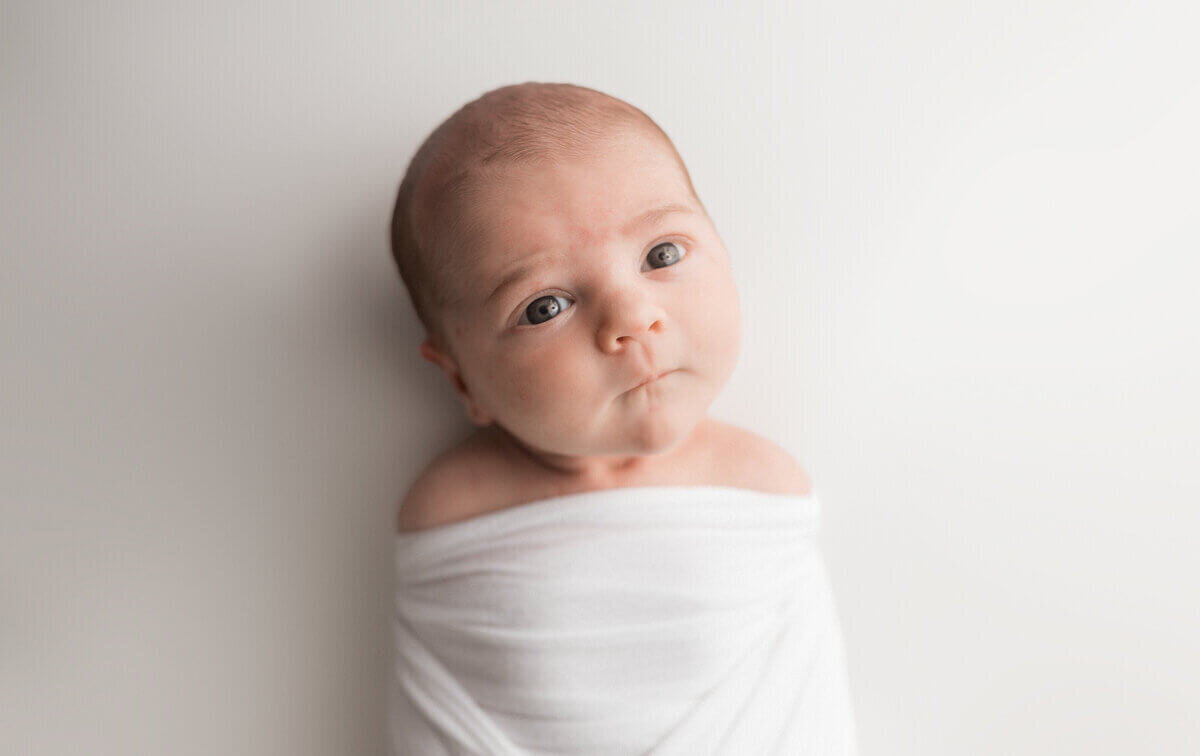 A newborn baby wrapped snugly in a white blanket, gazing wide-eyed at the camera, set against a minimalist white background.