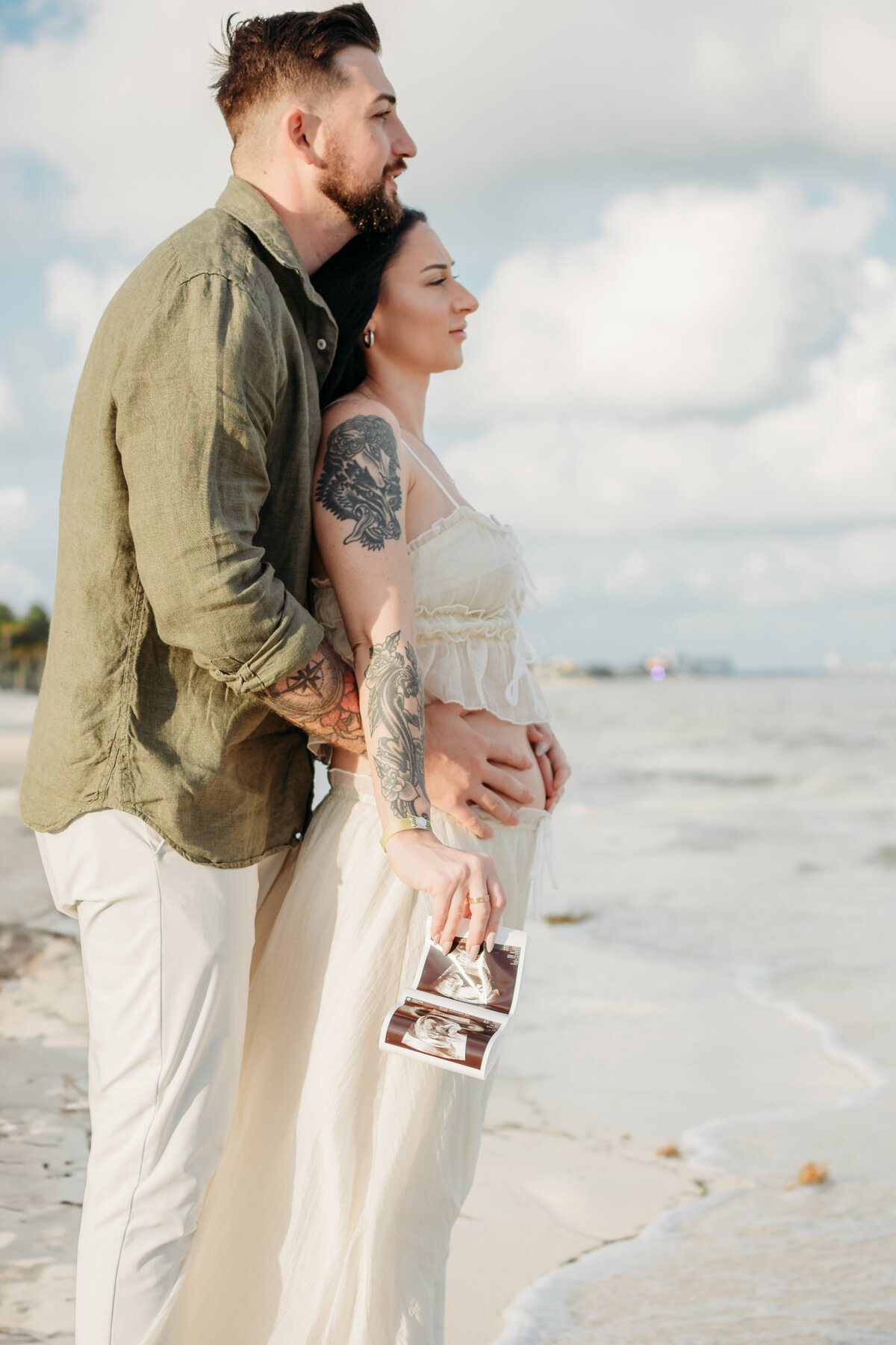 Soon to be mom and dad staring into the far distance of an ocean as they hold their baby ultrasound and embrace the love during a pregnancy announcement photoshoot