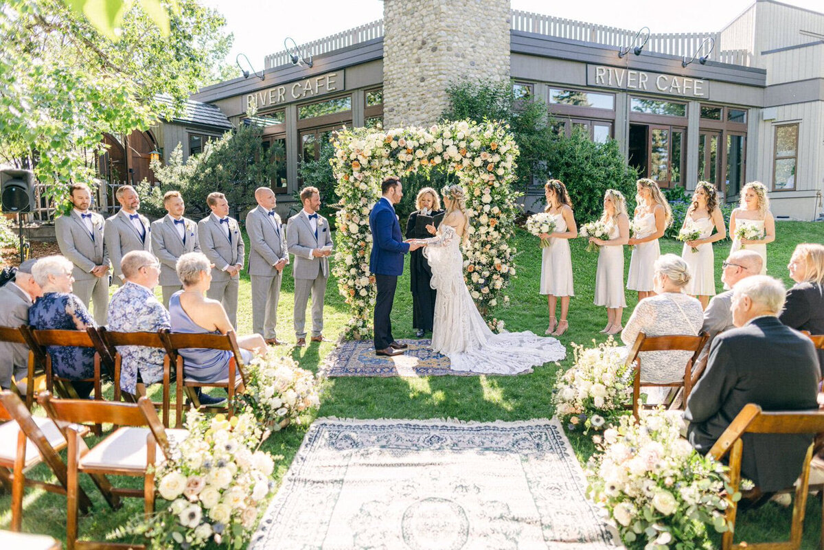 Outdoor wedding ceremony at River Café in Calgary, Alberta, featuring a couple exchanging vows under a vibrant floral arch. Surrounded by lush greenery, the bride in a flowing white gown and the groom in a classic black suit stand before their officiant. Bridesmaids in deep green dresses and groomsmen in black suits add to the elegant scene, while guests watch from floral-adorned wooden chairs. A romantic and picturesque setting along the Bow River.
