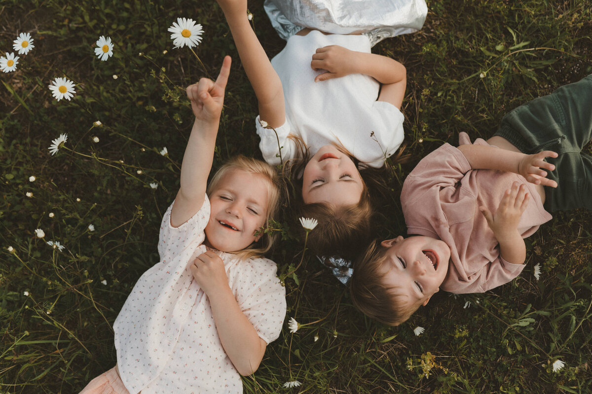 Foto van drie kinderen die tussen de bloemen liggen buiten en naar de wolken kijken.
