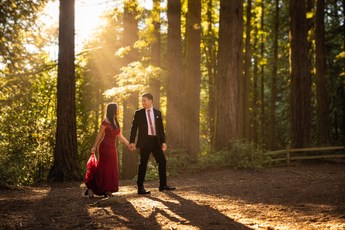 Engaged couple walks through forest with sun rays shining on them. They look at each other and smile. Photo by wedding photograph from Sacramento Philippe Studio Pro.