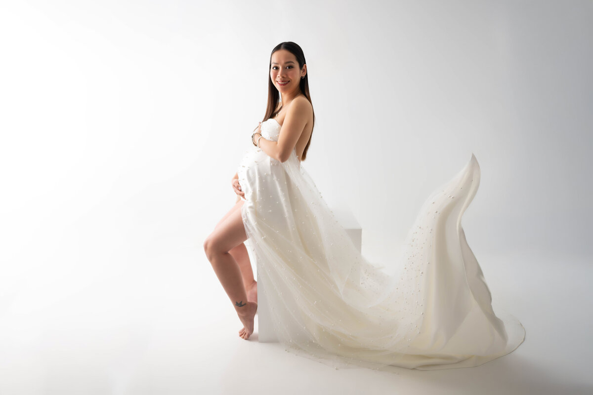 Pregnant women sitting on a posing box is dressed in a flowy white pearl embellished fabric. 