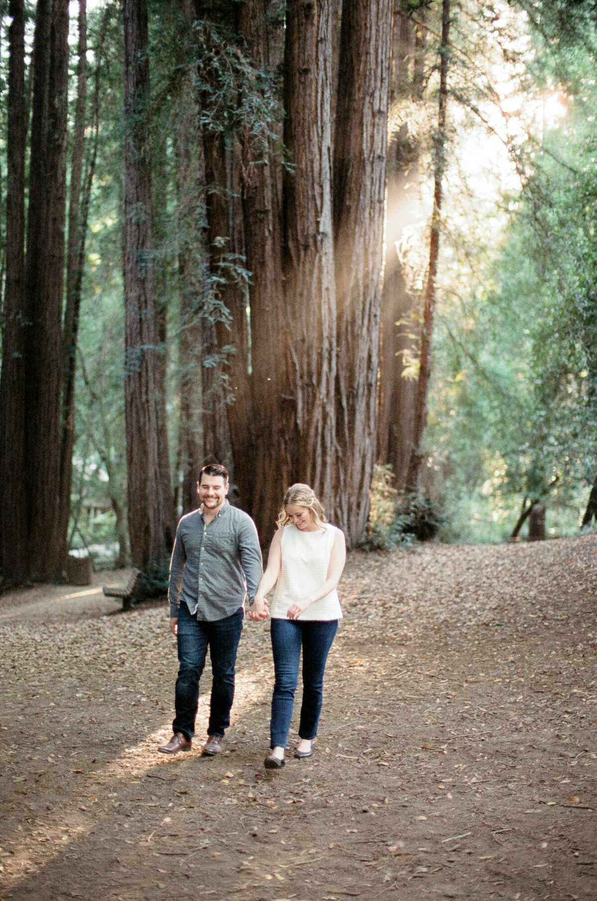 A loving couple hold hands and walk through tall trees with shimmering golden sunrays radiating through the woods.