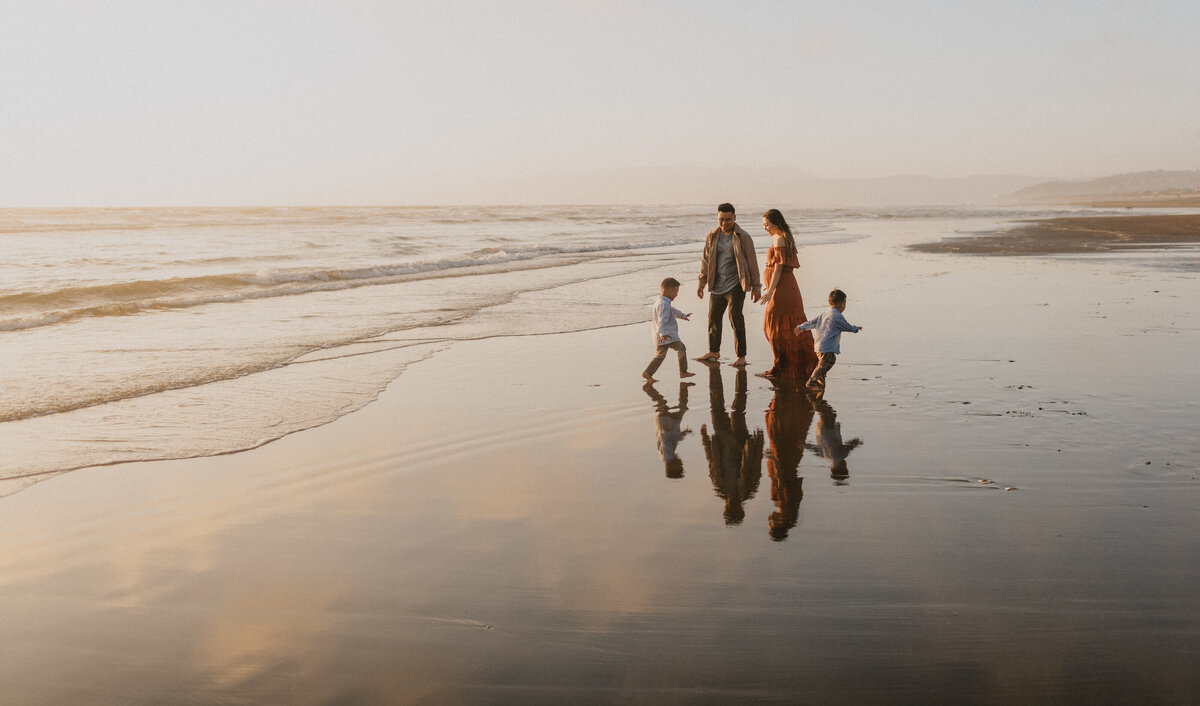 A family of four walks along a beach at sunset, their reflections visible on the wet sand as gentle waves roll in. two adults and two children enjoy the serene, golden-lit landscape in a family photoshoot