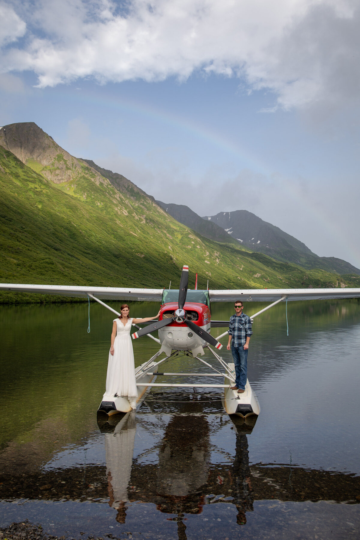A bride and groom stand on either side of a float plane while it is in the middle of a lake in Alaska.